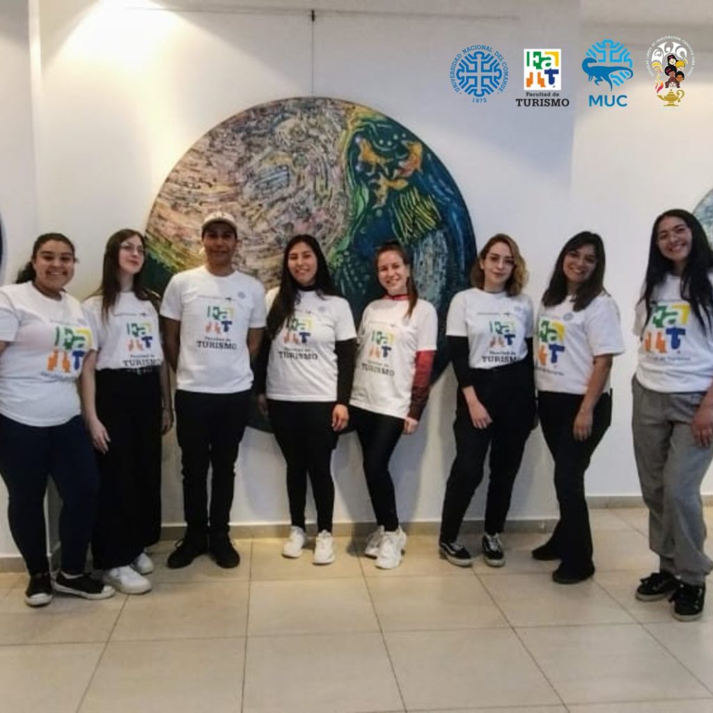 Grupo de estudiantes de la Facultad de Turismo posando frente a un mural circular. Visten camisetas blancas con el logotipo de la Facultad de Turismo. En la parte superior derecha se encuentran los logotipos de la Universidad Nacional del Comahue, la Facultad de Turismo y el MUC.